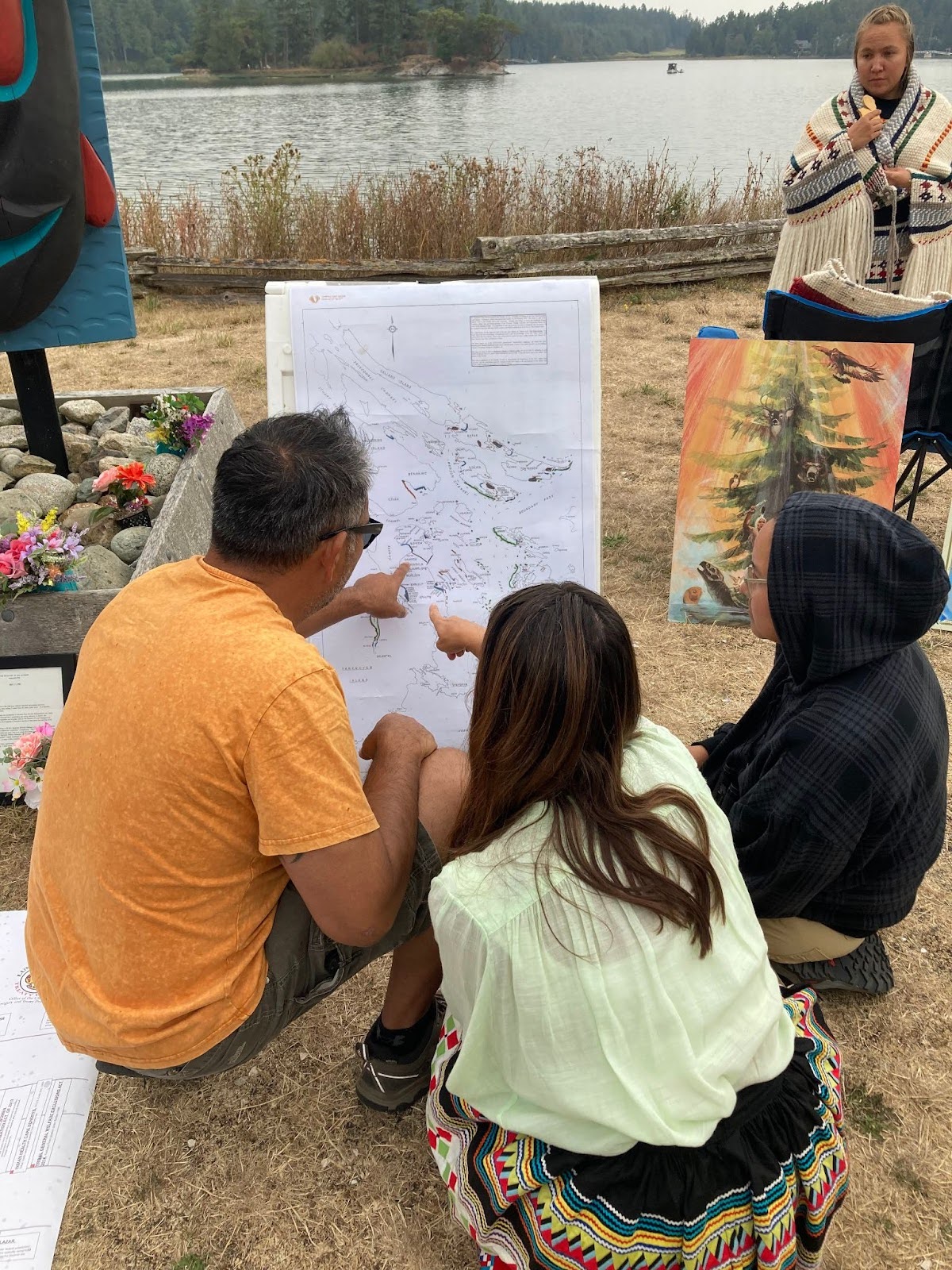 Three people sit in front of a large map printed on white paper, while two of them point at different areas
on the map. In the background a person wearing a Coast Salish woven shawl looks on while water and trees across
the water can be seen in the distance.